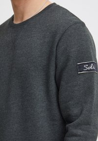 Donkergrijze sweatshirt met een ronde hals, zachte textuur en een patch op de linkermouw met het label "Soli." Regular fit met minimale ontwerpelementen.
