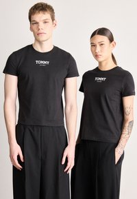 T-shirts pretas de algodão com mangas curtas e gola redonda, com o logótipo branco "TOMMY JEANS" no peito. Combinadas com calças pretas.