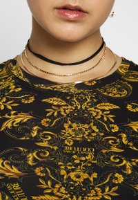 Svart blommig choker med en texturerad rem, lager på lager med två tunna guldkedjor, som kompletterar en mörk skjorta med gula blommönster.