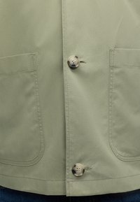 Veste verte olive claire à l'avant avec deux grandes poches plaquées et deux boutons ronds marbrés fixés au centre.