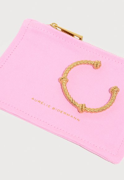 Ροζ υφασμάτινο πουγκί με χρυστό φερμουάρ, χρυσή στριφογυριστή λεπτομέρεια σε σχήμα "C" και την επιγραφή «Aurélie Bidermann» σε χρυσά γράμματα από κάτω.