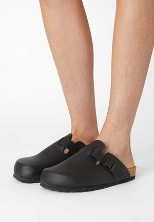 Black clogs with a rounded toe, two strap design, and side buckle accent. Featuring a cork footbed and textured rubber sole.