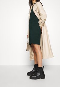 Beige trench coat kerroksittuna tummanvihreän sivusaumallisen mekon päälle. Mallilla on päällään mustat paksupohjaiset nilkkasukat ja hän pitää kädessään pientä, teksturoitua laukkua.