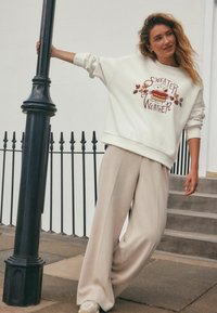 Kremowy sweatshirt z grafiką "Sweater Weather", zestawiony z beżowymi spodniami o szerokich nogawkach i białymi sneakersami. Teksturowana tkanina i luźny krój.