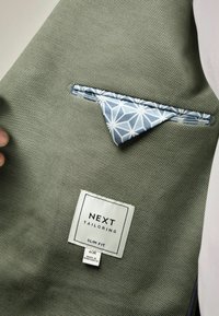 Oliwkowa, dopasowana marynarka z teksturowanym splotem, z jasnoniebieską poszetką w geometryczny wzór oraz metką z napisem "NEXT TAILORING."