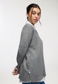 Maglione grigio a maniche lunghe con vestibilità ampia, dotato di scollatura a V e tre bottoni decorativi sul lato, indossato sopra una camicia bianca.