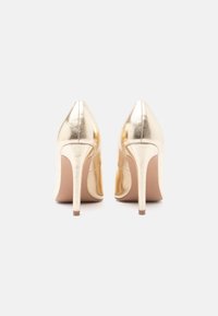 Um par de sapatos de salto alto dourados e brilhantes visto de costas sobre um fundo branco simples.