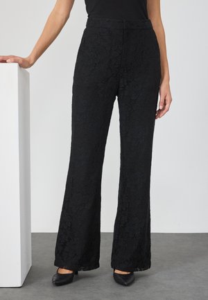 Pantalones de encaje negro con un patrón floral, con un diseño acampanado y una textura suave. Combinados con una blusa negra ajustada y zapatos negros.