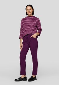 Lila, taillierte Hose kombiniert mit einem geometrischen, burgunderfarbenen Oberteil mit Dreiviertelärmeln und einem hohen Kragen. Glänzende schwarze Loafer.