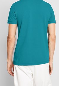 Teal kortärmad skjorta i ett mjukt, slätt tyg med rund hals och avslappnad passform, visad bakifrån, med synliga sömnadsdetaljer.