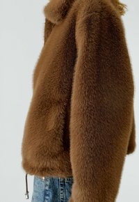 Bruine fuzzy cropped jacket gedragen met blauwe denim jeans tegen een effen lichte achtergrond, met een gedeeltelijk zijaanzicht.