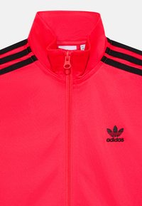 Raudona zip-up sportinė striukė su aukštu apykakle, dekoruota juodomis trijų juostų detalėmis ant pečių ir mažu juodu logotipu ant krūtinės.