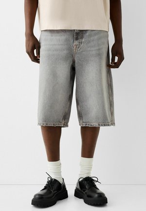 Homme portant un short en jean gris clair jusqu'aux genoux, des chaussettes blanches et des chaussures noires à lacets, se tenant contre un fond clair uni.
