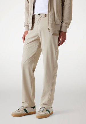 Beige trousers with side pockets, made of lightweight fabric. Paired with grey and green sneakers featuring a textured sole and branding.