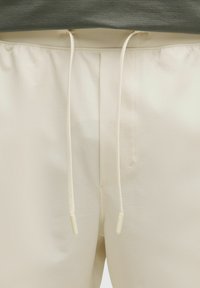 Pantaloni bej, cu croi lejer, confecționați dintr-un material texturat, având un brâu cu șnur cu vârfuri albe și un design minimalist.