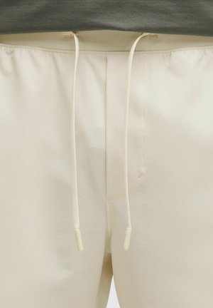 Pantalons beige à coupe décontractée en tissu texturé, dotés d'une taille élastique avec cordon de serrage et embouts blancs, au design minimaliste.