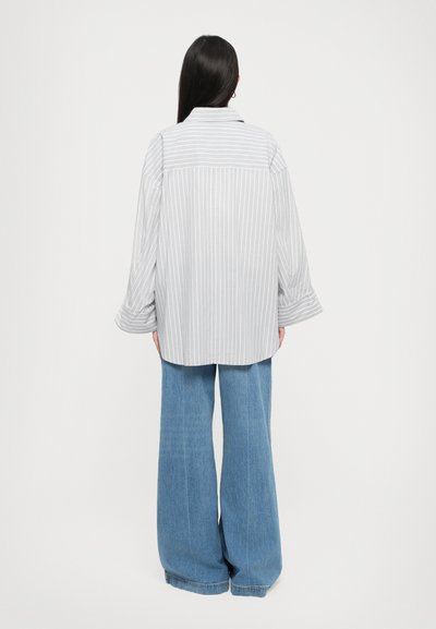 Woman with long dark hair wearing a loose gray and white striped shirt and wide-leg blue jeans, viewed from the back on a white background.