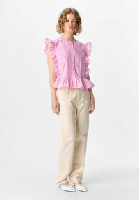 Blusa rosa con maniche a volant, chiusura con bottoni e orlo a peplum, abbinata a pantaloni a gamba larga color crema e scarpe bianche.