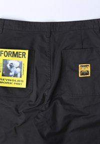 Zwarte werkbroek met een gemiddelde stoftextuur. Opvallende kenmerken zijn twee achterzakken, waarvan één met een gele label en grafics, en de andere met een geel embleem.
