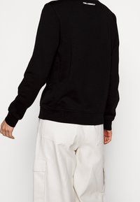 Sudadera negra de manga larga con puños acanalados. Presenta un cuello redondo y un logo en la parte trasera. Combinada con pantalones cargo de color claro.