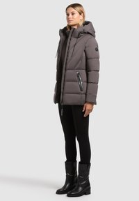 Naistearst kantakse halli vooderdatud kapuutsiga jope, musti retuuse ja musti põlvekõrguseid saapaid, seistes profiilis ühtse tausta ees.