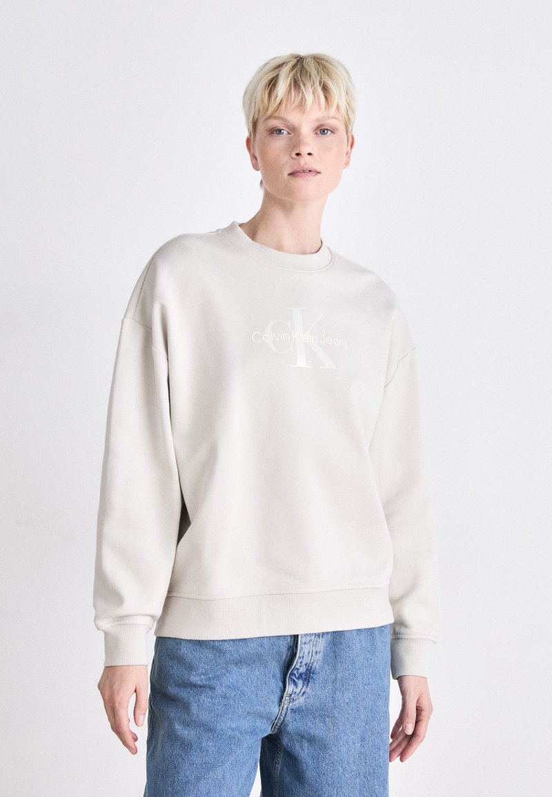 Light beige sweatshirt featuring a crew neckline and drop shoulders. Embroidered logo in the center. Paired with blue denim jeans.