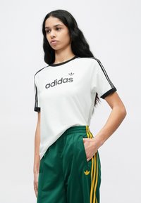 Biała koszulka z czarnym kołnierzykiem i logo Adidas, połączona z zielonymi spodniami sportowymi z żółtymi paskami i bocznymi kieszeniami.