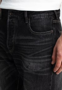 Nahaufnahme einer Person, die schwarze ausgewaschene Jeans und ein weißes Shirt trägt, mit der Hand an der Seite.