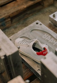 Baskets blanches avec languette rouge au talon étiquetée P448 reposant sur un moule à chaussure en métal dans un atelier.