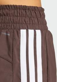 Shorts de sport marron avec une ceinture froncée, comportant trois bandes verticales blanches et une poche latérale en tissu texturé.