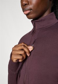 Persona con pelle scura che indossa una giacca a zip viola, mentre tira una piccola cerniera su una tasca sul petto vicino al colletto.