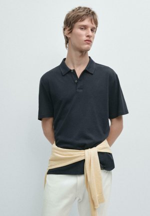 Junger Mann mit kurzen Haaren, der ein schwarzes Poloshirt, einen cremefarbenen Pullover um die Taille gebunden und weiße Hosen trägt, vor einem schlichten Hintergrund steht.