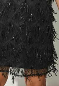 Mustat mekko, jossa on teksturoitu kangas, frangeja ja paljetteja. Helma on koristeltu mustilla helmiäisissä klustereilla ja paljeteilla.