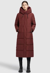 Abrigo acolchado burdeos con capucha, de diseño largo, con bolsillos laterales y textura acolchada. Combinado con botas negras hasta el tobillo.