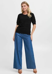 Haut noir texturé à manches courtes et ourlet festonné, associé à un pantalon en denim bleu à larges jambes et des escarpins bruns.