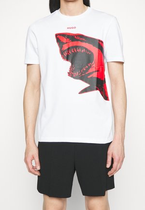 Mand iført en hvid T-shirt med en stor rød og sort hajgrafik og sorte shorts, stående mod en hvid baggrund.