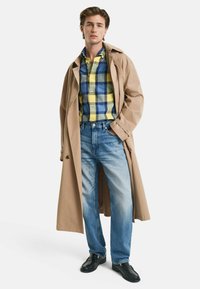 Beige Trenchcoat über einem blau-gelben Karobluse, kombiniert mit blassen blauen Jeans und schwarzen Schuhen. Das Outfit hat einen lässigen, schichten Look.