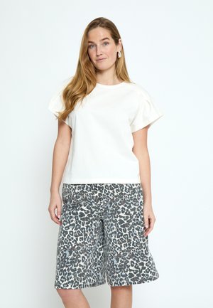 Vrouw met lang lichtbruin haar, gekleed in een wit topje met korte mouwen en losse, knie-lange shorts met luipaardprint, staand tegen een witte achtergrond.