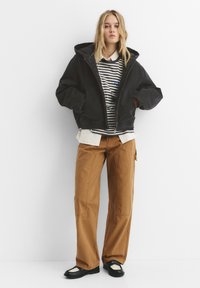 Jachetă neagră cu glugă peste un tricou cu mânecă lungă în dungi, cămașă bej stratificată, pantaloni maro cu picior larg și pantofi negri cu accente albe.