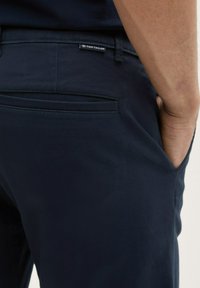 Pantaloni bleumarin dintr-un material moale, având un buzunar pe spate și un etichetă cu logo-ul Tom Tailor. Textură netedă cu o croială ajustată.