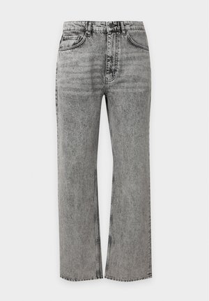 Jeune homme debout, une main dans la poche, portant une chemise blanche à manches longues, un jean gris ample et des baskets noires et blanches sur un fond uni.
