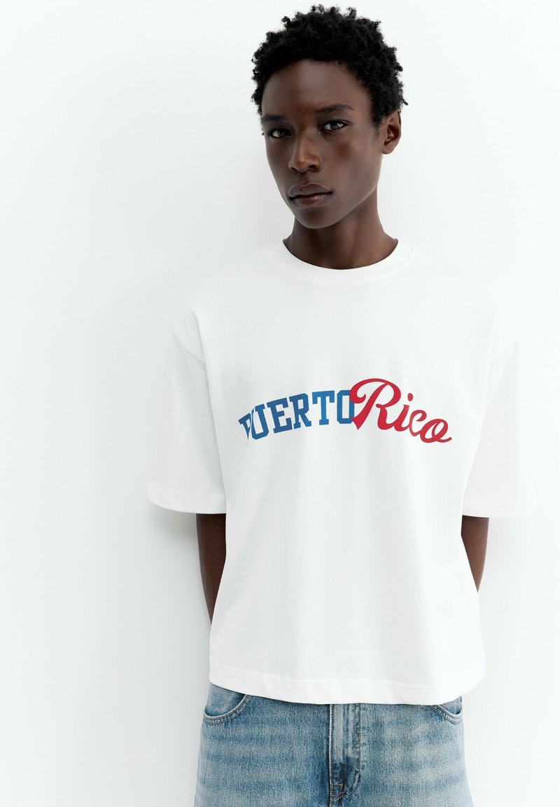 Jeune personne aux cheveux courts et bouclés portant un t-shirt blanc avec "Puerto Rico" en lettres bleues et rouges et un jean bleu sur fond blanc.