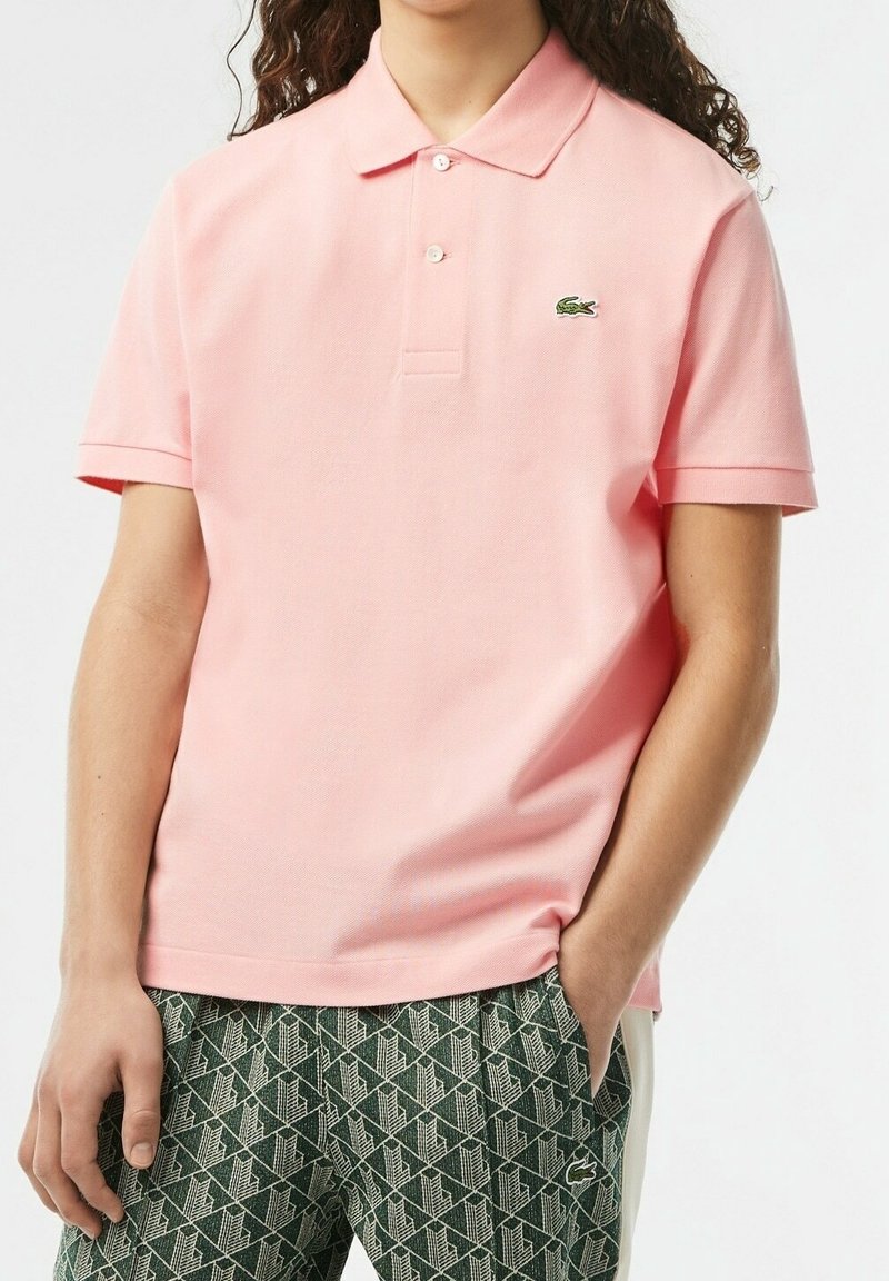 Polo à manches courtes de couleur rose clair, doté d'un col à trois boutons et du logo Lacoste. Associé à un bas vert à motifs.