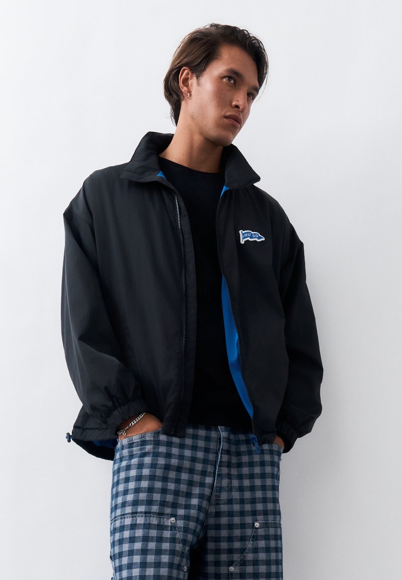 Jeune homme portant une veste noire zippée avec une doublure bleue et un pantalon à carreaux bleu-gris, debout les mains dans les poches devant un fond blanc.