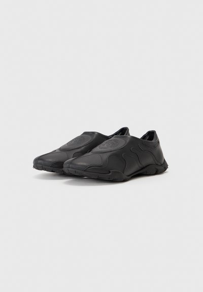 Zapatos negros tipo slip-on, fabricados con un material suave y un diseño moldeado. Presentan líneas curvas y una suela texturada para mayor agarre. Sin cordones ni cierres.