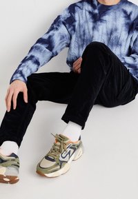 Niebiesko-biały sweter w dzianym wzorze tie-dye, czarne sztruksowe spodnie oraz zielone trampki z pomarańczowymi akcentami; siedząca pozycja z rozluźnioną postawą.