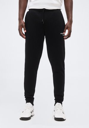 Άνδρας που φοράει μαύρο παντελόνι jogger με λευκά γράμματα στο μηρό και λευκά αθλητικά παπούτσια με μαύρες λεπτομέρειες, στέκεται σε απλό φόντο.