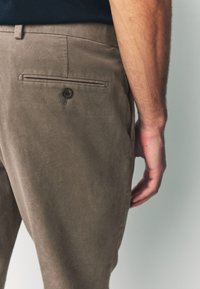 Pantalon kaki à texture lisse, doté d'une poche arrière avec fermeture à bouton, vu de côté avec une main posée à proximité.