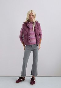 Joven con cabello rubio largo vistiendo un chaleco acolchado morado, un suéter rosa con estampado, unos vaqueros cortos grises y unas zapatillas burdeos.