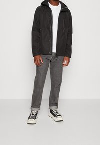 Zwarte lichtgewicht jas met capuchon, ritssluitingzak, wit T-shirt, grijze opgerolde spijkerbroek en zwarte sneakers met witte zolen.